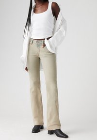 Jeans acampanados de un beige claro, hechos de denim, con un acabado desgastado, diseño de cinco bolsillos y herrajes de metal, combinados con una camiseta sin mangas blanca y una camisa de gran tamaño.
