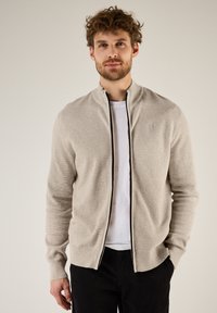 Chaqueta de punto beige con cremallera y cuello alto, que presenta un patrón texturizado y detalles en cremallera negra. Lleva puesta sobre una camiseta blanca y pantalones negros.