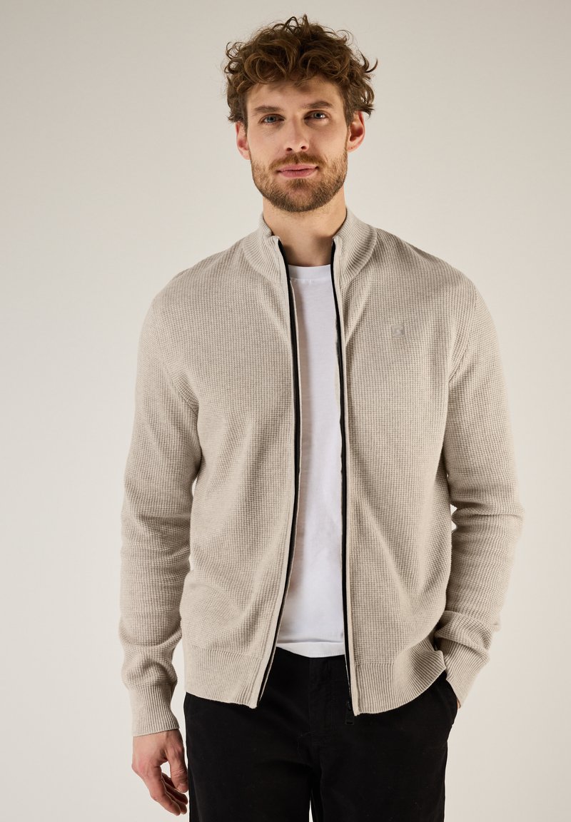 Chaqueta de punto beige con cremallera y cuello alto, que presenta un patrón texturizado y detalles en cremallera negra. Lleva puesta sobre una camiseta blanca y pantalones negros.