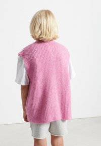 Ružičasta pletena brezrokavnik z teksturiranim rebrastim vzorcem, nosi se nad belo srajco, v kombinaciji s sivo kratkimi hlačami.