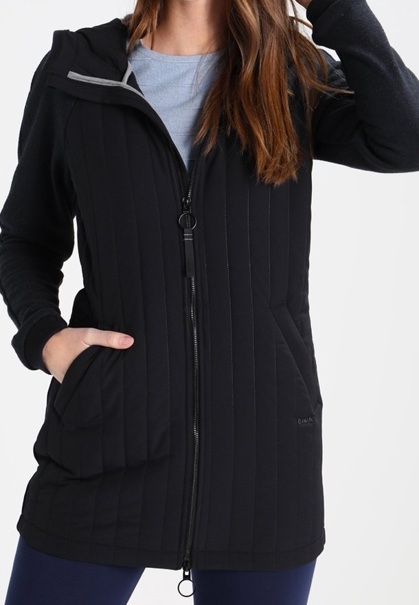 Chaqueta negra con capucha, textura acolchada, cierre con cremallera, dos bolsillos delanteros y forro interior gris, diseñada para brindar calor y comodidad.