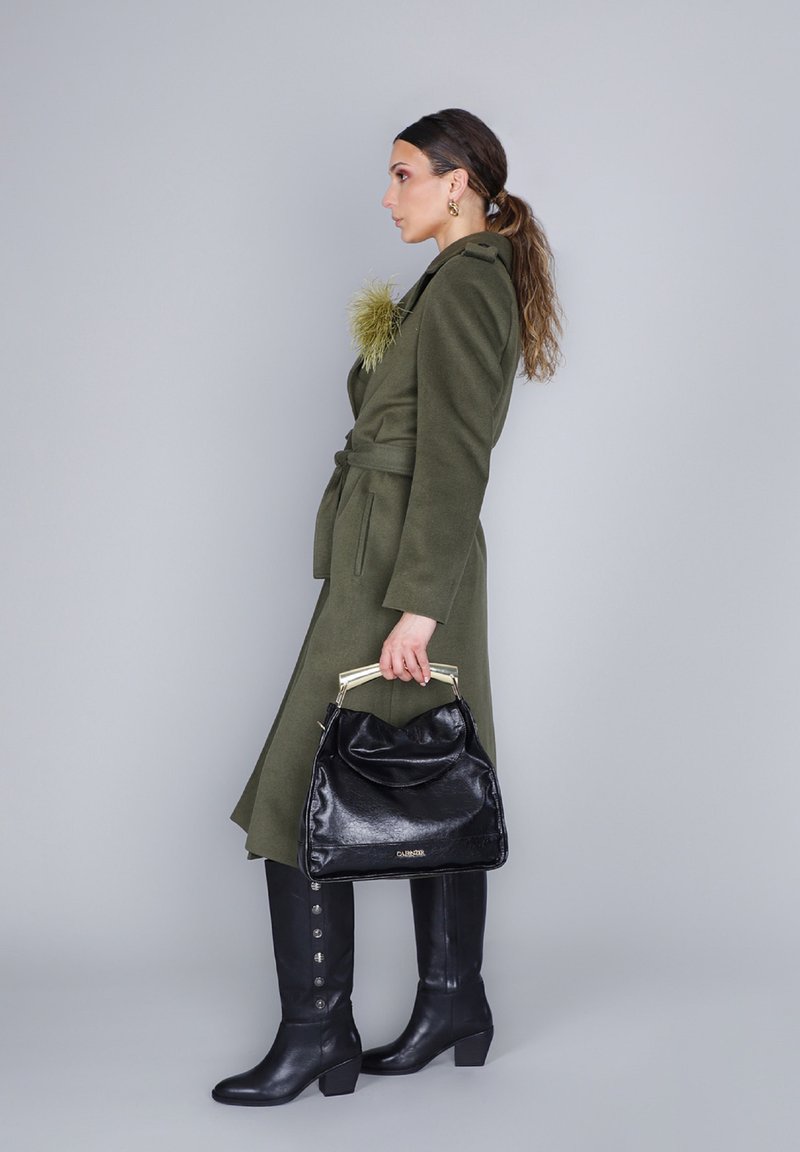 Oliivinvihreä takki, jossa on tekstuuriltaan turkista muistuttava yksityiskohta; musta käsilaukku kiiltävällä pinnalla; polven yli ulottuvat mustat saappaat sivunappien kanssa; neutraali tausta.