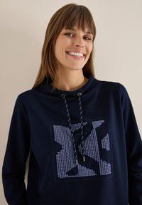 Sudadera con capucha de color azul marino que presenta un patrón geométrico en un azul más claro y acentos metálicos, con cordones y un ajuste relajado.