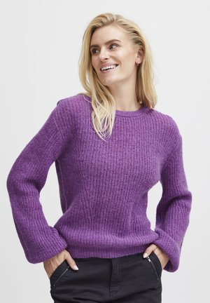 Purple knit sweater with textured fabric, round neckline, and wide sleeves. Worn with black pants, featuring a side zip pocket.