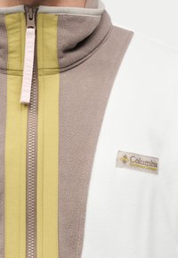 Κοντινό πλάνο ενός μπουφάν από φλις με μπεζ, κίτρινες και λευκές λεπτομέρειες και φερμουάρ μπροστά, που εμφανίζει το λογότυπο της Columbia Sportswear Company.