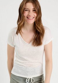 T-shirt blanc léger en V avec des manches courtes roulées et un motif texturé subtil. À associer avec un pantalon olive vert ample à cordon.