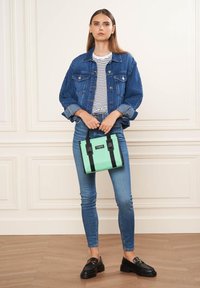 Sac à main vert menthe avec des poignées noires, présentant une fermeture éclair. Le modèle porte un jeans en denim, une chemise rayée et un jean bleu slim.