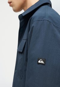 Πλάγια όψη ενός ατόμου που φοράει μπλε πουκάμισο με κουμπιά, με τσεπάκι με καπάκι και σήμα λογότυπο της μάρκας σε μαύρο και λευκό στο μανίκι.