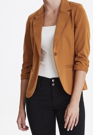 Femme portant un blazer moutarde aux manches retroussées, un débardeur blanc et un pantalon noir taille haute avec double boutons.