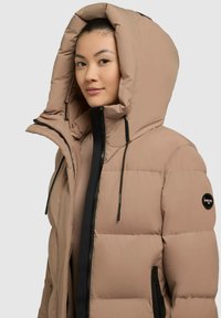 Doudoune beige avec une capuche oversize, fermeture éclair noire et cordons de serrage réglables. Comprend un patch rond en logo sur la manche gauche.