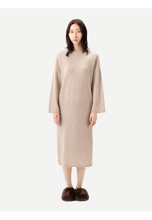 Frau steht und trägt ein langes beiges Pulloverkleid mit weiten Ärmeln und dunkelbraunen flauschigen Hausschuhen auf einfarbig hellem Hintergrund.