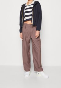 Granatowa kurtka z kapturem, krótka bluzka w paski i beżowe spodnie o szerokich nogawkach. Białe trampki na lekkiej platformie. Gładkie tło.