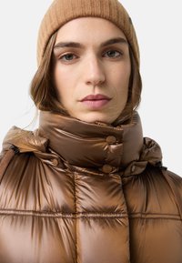 Braune wattierte Jacke mit hohem Kragen, glänzendem Finish und Knopfdetails. Kombiniert mit einer gerippten, hellbraunen Strickmütze.