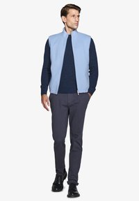 Gilet senza maniche di colore azzurro chiaro sopra un maglione blu scuro, abbinato a pantaloni grigi scuri e scarpe nere. Tessuto liscio con chiusura a zip.