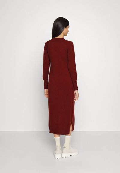 Vestido longo burgundy feito de material de malha suave, com mangas longas, um decote redondo canelado e aberturas laterais. Usado com botas brancas grossas.
