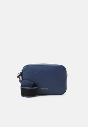 Sac bandoulière rectangulaire en cuir bleu marine avec une sangle noire ajustable, des ferrures dorées et un petit logo centré à l'avant.