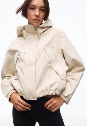Mladá žena ve světlé béžové bundě s kapucí, velkými kapsami a elastickým lemem stojí před bílým pozadím.