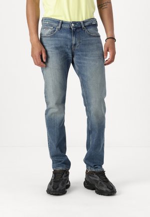 Personne portant un jean bleu délavé, des chaussures de sport noires et un t-shirt jaune clair visible de la taille vers le bas sur un fond blanc uni.