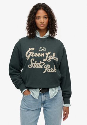 Mørkegrøn sweatshirt med hvid tekst "Green Lakes State Park." Båret over en lyse stribet skjorte. Casual pasform, ribbede manchetter og kant.