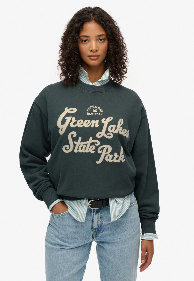 Sudadera verde oscuro con texto blanco "Parque Estatal Green Lakes". Llevada sobre una camiseta de rayas clara. Ajuste casual, con puños y dobladillo acanalados.
