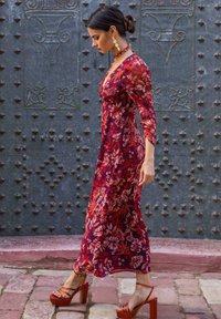 Langes Blumenkleid in Rottönen mit langen Ärmeln und V-Ausschnitt. Das Model trägt orangefarbene Plateau-Sandalen. Der Hintergrund zeigt eine strukturiert metallische Tür.