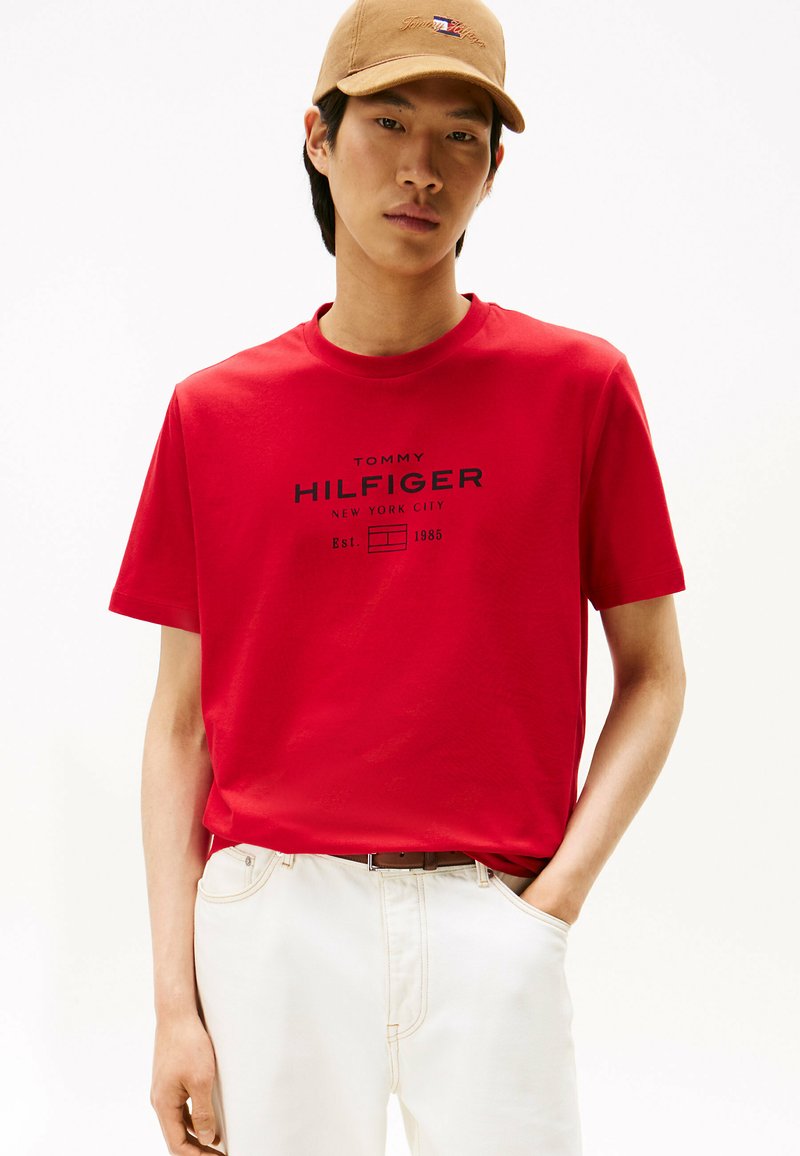 Rød T-shirt med korte ærmer, der har et sort logo-print med teksten "TOMMY HILFIGER" på forsiden; iført med lyse jeans og en tan farvet kasket.