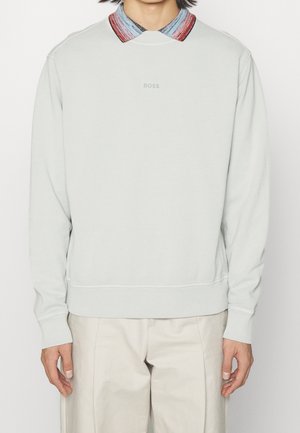 Person trägt ein hellgraues Sweatshirt mit dem "BOSS"-Logo mittig auf der Brust, darüber ein mehrfarbig gestreiftes Hemd mit Kragen und beigefarbene Hosen.