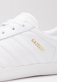 White leather sneaker featuring diagonal ribbed texture, gold "Gazelle" branding, and a rubber sole with textured detailing.
