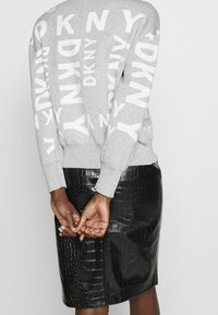 Šedý svetr s velkým bílým potiskem loga "DKNY", doplněný o černou sukně z umělé krokodýlí kůže. Texturované materiály a přiléhavý střih.