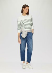Gestreifter Langarmpullover in Weiß und Grün, kombiniert mit lockeren blauen Denimhosen und weißen Sneakern mit strukturiertem Sohlen. Grauer Strickpullover um die Taille getragen.