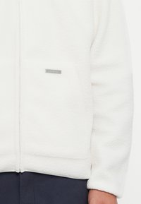White fleece jacket with a zip, featuring a front pocket and a small silver logo tag, soft texture, and relaxed fit design.