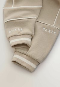 Bežové manžety mikiny s bielymi akcentmi, s výšivkou "BAKER" v bielej farbe. Tkanina je mäkká s rebrovanou textúrou a pruhovanými detailmi.