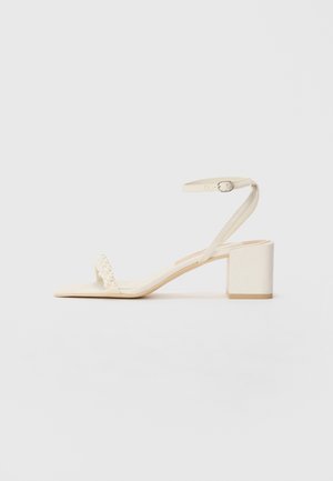 Sandalo con tacco blocco beige realizzato in materiale liscio, dotato di una sottile cinghia alla caviglia e di una cinghia per le dita decorata con perle. Sole piatto.