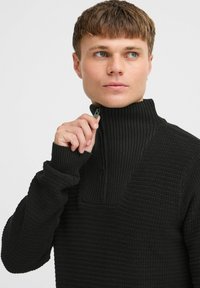 Pulover negru din tricot, cu guler înalt, textură ribbed și detaliu cu fermoar la gât, având mâneci lungi și o croială lejeră.