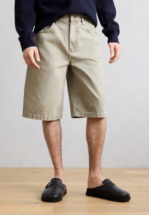 Persona che indossa pantaloncini beige fino al ginocchio, scarpe nere slip-on e un maglione blu navy a maniche lunghe, in piedi su un pavimento in legno.
