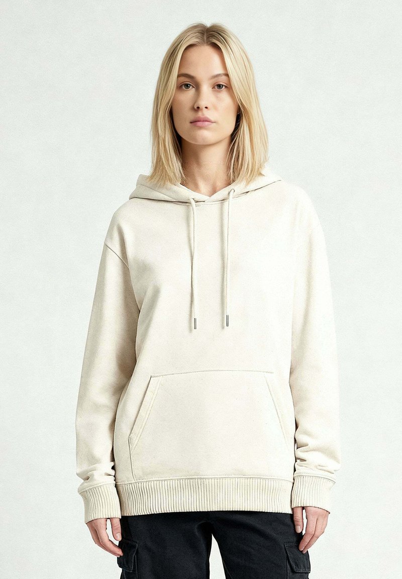 Femme blonde portant un sweat à capuche beige clair avec une poche avant et un pantalon cargo noir, debout devant un fond clair uni.