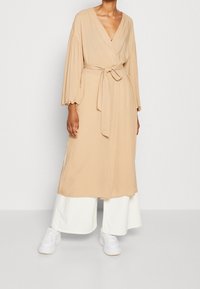 Beżowa sukienka w formie owinięcia z długimi bufkami, paskiem w talii i bocznymi rozcięciami, zestawiona z białymi spodniami o szerokich nogawkach i białymi trampkami.