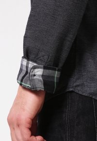 Grå denimskjorteærme med rullet ternet manchet i sort, hvid og grønne farver. Bemærkelsesværdig fiskebensstruktur på det grå stof.