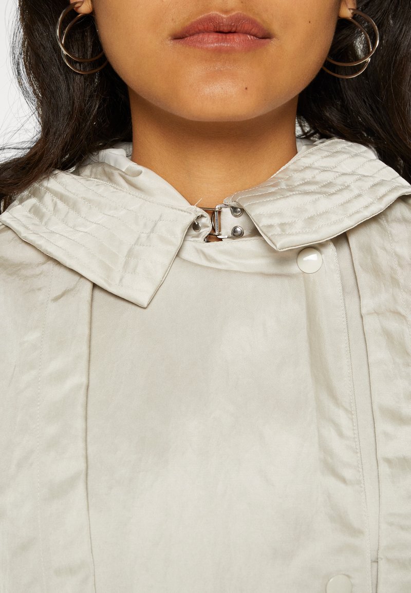 Chaqueta gris claro con capucha texturizada, que cuenta con un cierre de botones y detalles metálicos. Tejido suave y semibrillante con un diseño ajustado.