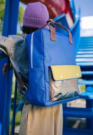 Enfant portant un bonnet tricoté violet et un t-shirt gris porte un sac à dos bleu avec une poche avant jaune et argentée, montant des escaliers bleus en plein air.