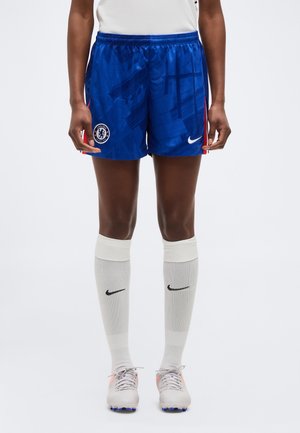 Persona vistiendo pantalones cortos de fútbol Chelsea azules, calcetas blancas de Nike hasta la rodilla y zapatos de fútbol blancos, parada frente a un fondo liso.