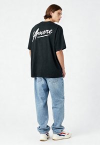 Juodas per didelis marškinėliai su baltais "Amore" tekstu ant nugaros, derinami su šviesiai mėlynomis plačiomis džinsomis ir baltais sportbačiais su raudonais ir mėlynais akcentais.