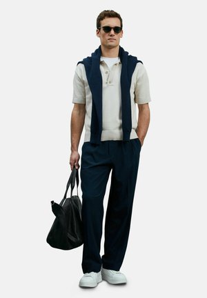 Witte polo met korte mouwen en kraag, marineblauwe trui over de schouders, marineblauwe broek en witte sneakers; houdt een zwarte tas vast.