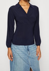 Marinblå blus med lång ärm och v-ringning, sammanknuten detalj vid midjan och mjukt, slätt tyg. Matcher med en ljus denimkjol.