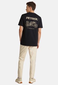 Camiseta de algodón negra con un gráfico de motocicleta en color crema en la espalda, combinada con pantalones claros y zapatillas deportivas.