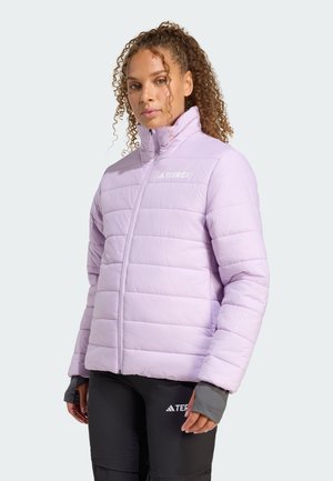 Una donna con capelli ricci indossa un giubbotto imbottito di un viola chiaro e pantaloni neri con un logo, il tutto su uno sfondo chiaro.