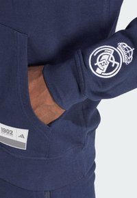 Marinblå hoodie i texturerad tyg, med en logga på ärmen, en framficka och en etikett som visar "1902 Real Madrid" och Adidas-logotyp.