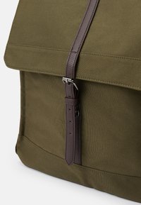 Sac en tissu vert olive avec un rabat pliable, une bandoulière en cuir brun et une boucle en argent. Surface texturée avec des détails de couture visibles.