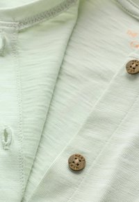 Tissu tricoté vert clair avec des boutons marron gravés "NEXT" et des boutonnières visibles sur le vêtement.