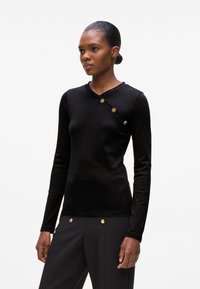 Maglietta nera a maniche lunghe con scollo a V e bottoni laterali diagonali in oro. Realizzata in un tessuto liscio con un design aderente.